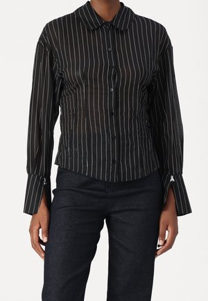 Sort langærmet skjorte med knapper og hvide lodrette nålestriber, båret med mørke bukser, med forlængede manchetter og lynlåsdetailler.