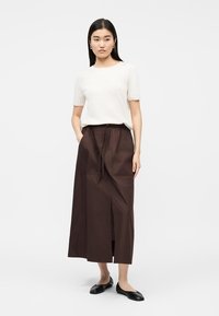 Femme portant un haut blanc à manches courtes, une jupe midi marron fendue à l'avant avec cordon de serrage, et des ballerines noires, debout les mains dans les poches.