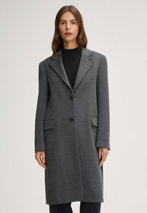 Vrouw draagt een lange, donkergrijze wollen jas over een zwart topje, staand tegen een effen lichte achtergrond.