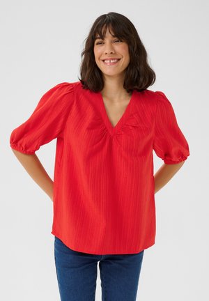 Blusa rossa a maniche a sbuffo con scollatura a V, caratterizzata da strisce verticali testurizzate e una vestibilità rilassata, abbinata a jeans blu.