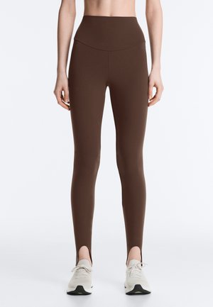 Frau trägt braune Leggings mit hoher Taille und Knöchelschlitzen, kombiniert mit weißen Sportschuhen, steht vor einem schlichten Hintergrund.