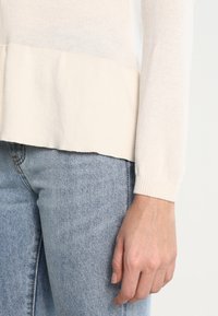 Jasny beżowy sweter z dzianiny z falbanką u dołu. Tekstura jest miękka i lekko przejrzysta. Sweter ma długie rękawy i prążkowane mankiety.
