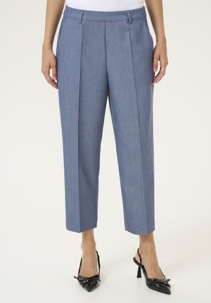 Pantaloni su misura azzurro chiaro con pieghe frontali, lunghezza alla caviglia e una texture liscia. Indossati con scarpe nere con tacco e punte appuntite.