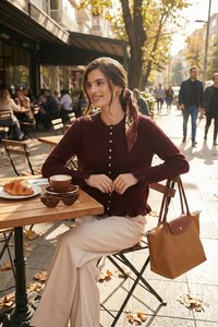 Femme en pull bordeaux et pantalon beige assise à une table de café en terrasse avec un café, un croissant et des lunettes de soleil par une journée d'automne ensoleillée.