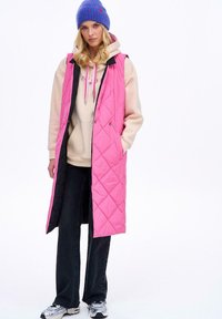 Kurtka pikowana w odcieniach różu i czerni, wykonana z gładkiego materiału. Nosi się ją na beżowej bluzie z różowymi sznurkami, w połączeniu z ciemnymi dżinsami i trampkami.