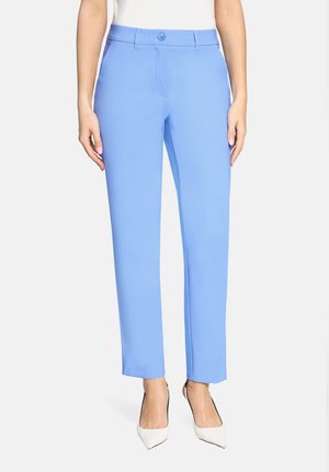 Frau trägt schlanke hellblaue Hosen und weiße spitze Absatzschuhe, steht vor einem einfarbigen hellen Hintergrund.