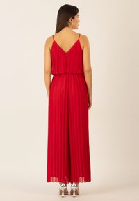 Roter, plissierter Jumpsuit mit V-Ausschnitt, dünnen Trägern und einem geschichteten Oberteil. Mit einem Reißverschluss und fließendem Design, kombiniert mit silbernen High Heels.