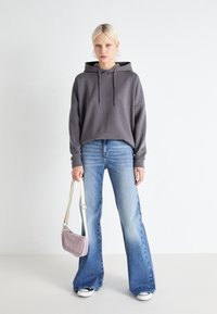 Grå hettegenser med snøring, lyseblå vide jeans og en liten lilla veske med en beige stropp; svart og hvite sneakers på føttene.
