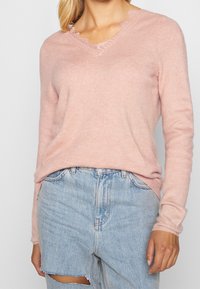 Rosa V-Ausschnitt-Pullover mit strukturiertem Finish und ausgefransten Kanten, kombiniert mit leicht verwaschenen hellblauen Jeans.
