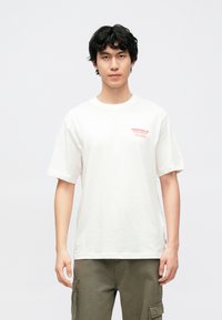 Hvid bomuld T-shirt med korte ærmer, afslappet pasform, med rød tekst på venstre bryst og rund halsudskæring, båret med grønne bukser.