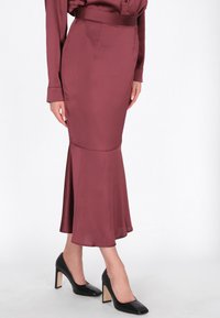 Kaštanová saténová mořská panna sukně s přiléhavým pasem a rozšířeným lemem. Nošena s černými lodičkami na vysokém podpatku, hladká textura a minimální design.