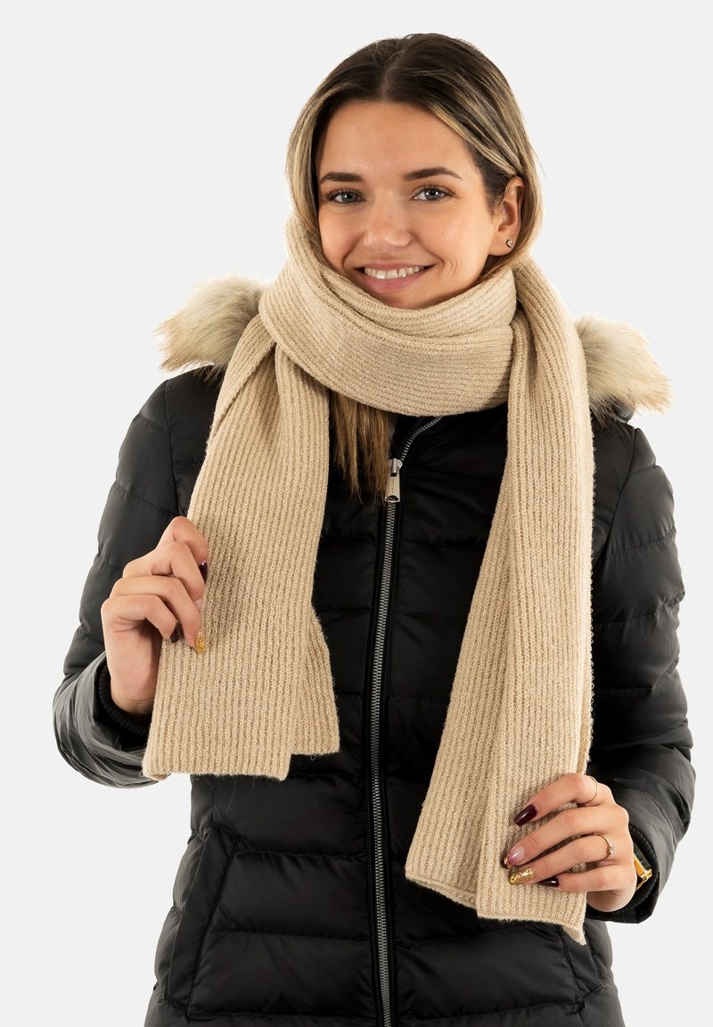 Beige geribbelde gebreide sjaal, gemaakt van zacht materiaal, met een chunky textuur, vastgehouden door een persoon die een zwarte pufferjack draagt.