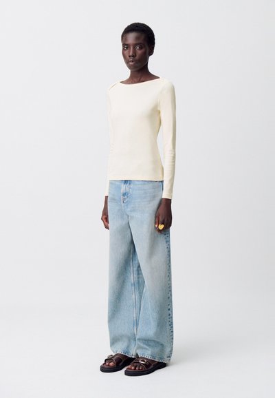 Donna in piedi con una maglietta a maniche lunghe crema, jeans ampi blu chiaro, sandali neri con fibbia e un grande anello d'oro sulla mano destra.