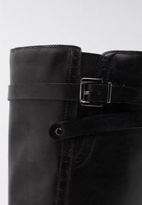 Bota de cuero negra con la parte superior redondeada, que presenta una correa con hebilla y un panel lateral elástico. Textura suave con costuras sutiles y detalles perforados.