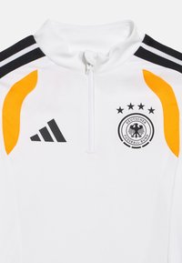 Balta sporta jaka ar melniem un oranžiem akcentiem, Adidas logotips kreisajā krūtīs un Vācijas futbola komandas emblēma ar četriem zvaigznītēm labajā krūtīs.