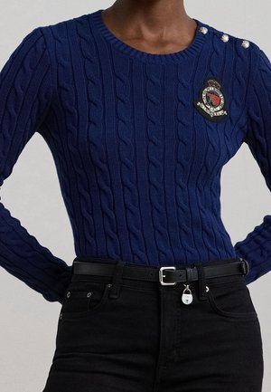 Henkilö, jolla on päällään tummansininen kaapelikuvioitu neulepaita, jossa on napit ja kirjailtu vaakuna, yhdistettynä mustiin farkkuihin ja vyöhön, jossa on pieni lukonmuotoinen riipus.