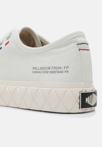 Baskets en toile blanche avec semelle en caoutchouc texturée, coutures rouges et bleues au talon et étiquette de la marque Palladium sur le côté et le talon.