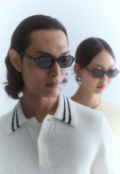 Lunettes de soleil noires avec des verres rectangulaires, portées avec un polo blanc texturé avec un col rayé. Une deuxième silhouette dans un haut clair est floue à l'arrière-plan.
