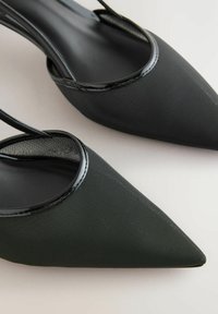 Nahaufnahme von schwarzen Slingback-Schuhen mit spitzer Zehenform aus strukturiertem Stoff und glänzenden Details auf einer hellen Fläche.