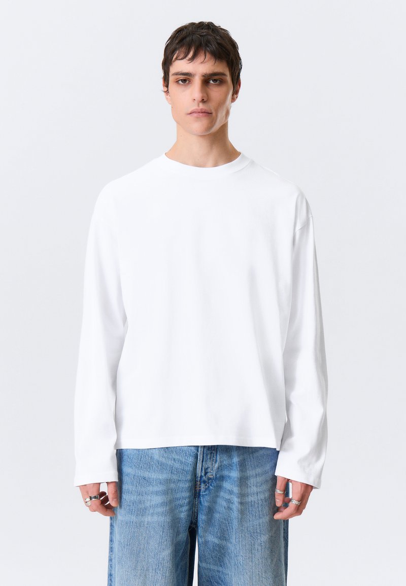 Hvit skjorte med lange ermer og rund hals, løs passform og glatt tekstur, kombinert med lyseblå vide bukser.