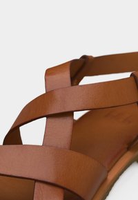 Καφέ δερμάτινο σανδάλι με φαρδιές διασταυρούμενες λουρίδες και επίπεδη σόλα σε λευκό φόντο.