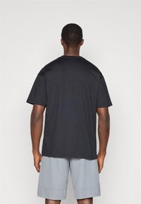 Μαύρη κοντομάνικη μπλούζα, από βαμβακερό υλικό, άνετη εφαρμογή, με στρογγυλή λαιμόκοψη, ορατές λεπτομέρειες ραφής, φορεμένη με γκρι αθλητικά σορτς.