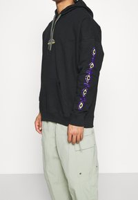 Felpa nera in cotone con tasca frontale, caratterizzata da un motivo decorativo in viola e giallo lungo la manica sinistra.