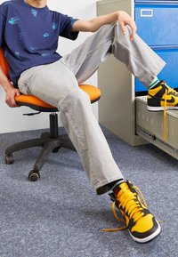 Nike t-shirt in navy with blue logos, light gray straight-leg jeans, and black/yellow sneakers with orange laces, seated on an orange chair.