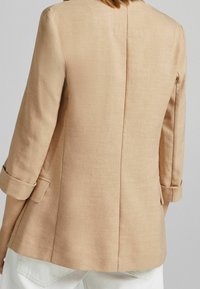Blazer en lin beige avec une coupe ajustée, des revers crantés, des manches retroussées et un détail de couture au dos. Texture lisse et design minimaliste.