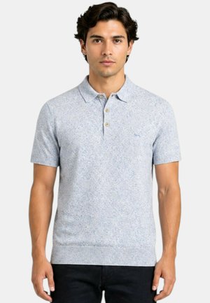 Giovane uomo con capelli scuri che indossa una polo azzurro chiaro a maniche corte con un motivo floreale sottile e pantaloni neri.