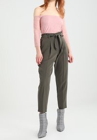 Top in pizzo rosa con spalle scoperte abbinato a pantaloni a vita alta verde oliva con fiocco. Stivaletti alla caviglia a quadri grigi completano il look.