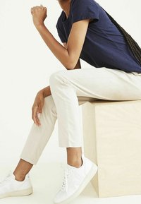 Marinblå kortärmad t-shirt, krämfärgade slim-fit byxor och vita sneakers med strukturerade sulor. Modell som sitter på en ljus träkub.