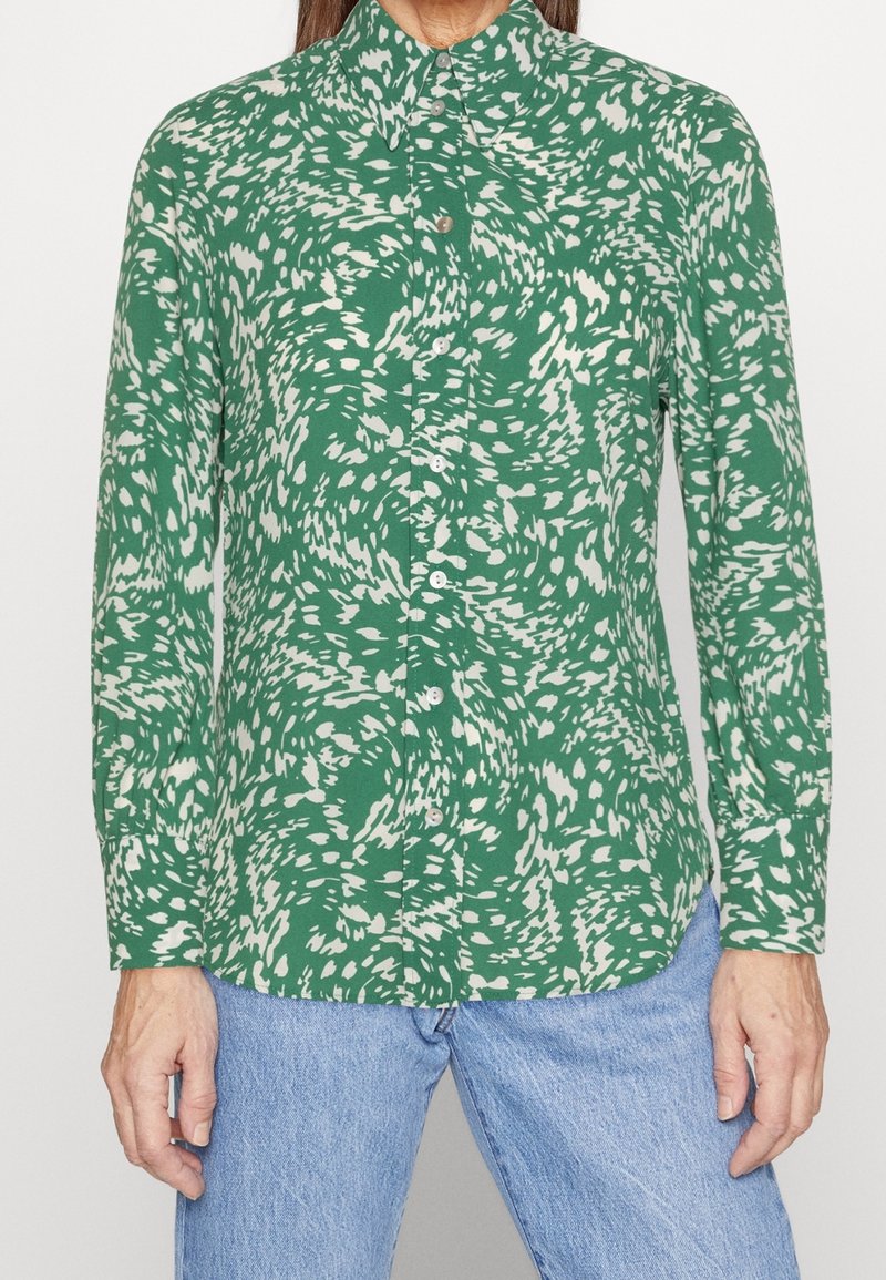 Chemise verte à boutons avec un motif blanc tourbillonnant, col chemise et manches longues, portée avec un jean bleu clair.