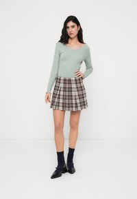 Φωτεινό πράσινο ριμπ μακρυμάνικο τοπ συνδυασμένο με καφέ και λευκή καρό φούστα καμπάνα. Η μοντέλο φοράει μπλε ναυτικά ριμπ κάλτσες και μαύρα μυτερά παπούτσια.