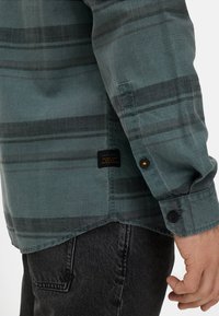 Türkis kariertes Hemd mit schwarzen Streifen, Knopfmanschetten und einem Stoffetikett am Saum. Die Textur wirkt glatt und eignet sich für legere Kleidung.