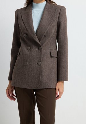 Frau trägt einen braunen doppelt geknöpften Fischgrät-Blazer über einem hellblauen Strickrollkragen und braune Hose, stehend vor einem schlichten Hintergrund.