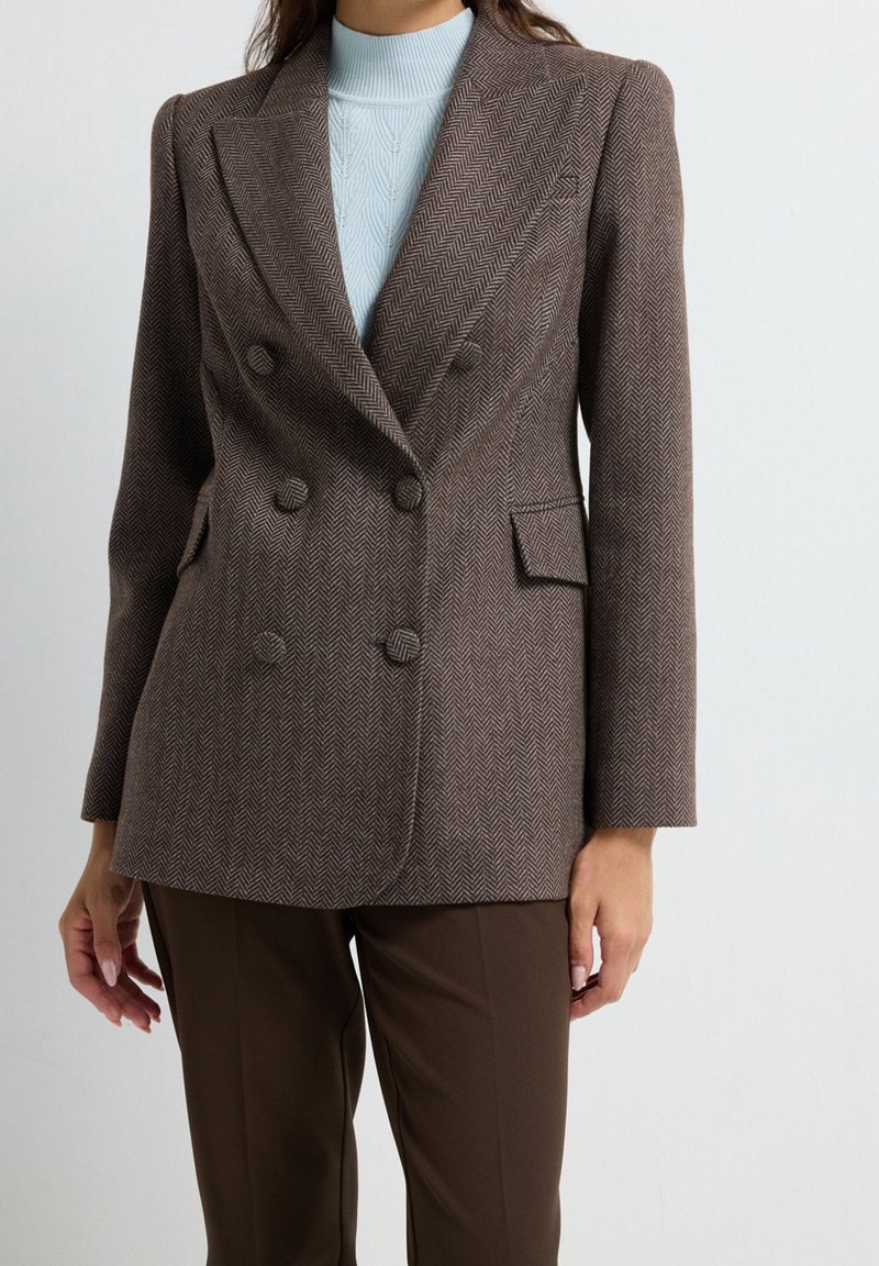 Femme portant un blazer croisé marron en chevrons sur un pull à col roulé tricoté bleu clair et un pantalon marron, debout devant un fond uni.