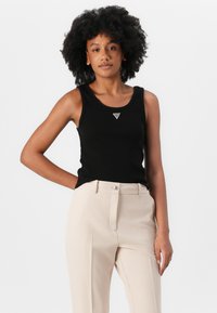 Black tank top with a rounded neckline, featuring a small silver triangular logo. Worn with light beige tailored trousers. Smooth fabric texture.
