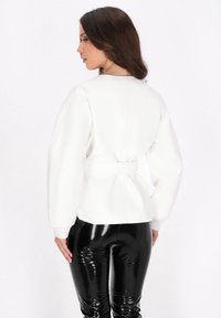 Weißer Pullover mit geraffter Taille und gerippten Bündchen, kombiniert mit glänzenden schwarzen Leggings. Glatte Textur und maßgeschneiderter Schnitt.