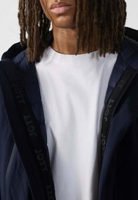 Persona con trecce indossante una camicia bianca e una giacca blu navy con fodera interna nera e fasce nere con marchio.