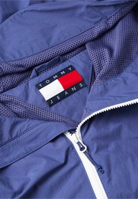 Gros plan d'une veste bleue avec une fermeture éclair blanche et une doublure en mesh, montrant le patch du logo Tommy Jeans en rouge, blanc et bleu marine.