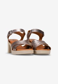 Sandalias de cuero trenzado bronce con tacón bloque, con correas de tobillo ajustables y una plantilla suave color marrón claro, sobre una suela de color claro.