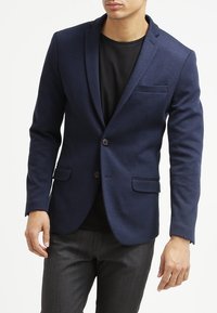 Marinblazer tillverkad av texturerad tyg, med snibbkrage, enkel knappstängning och en bröstficka, parat med en svart t-shirt.