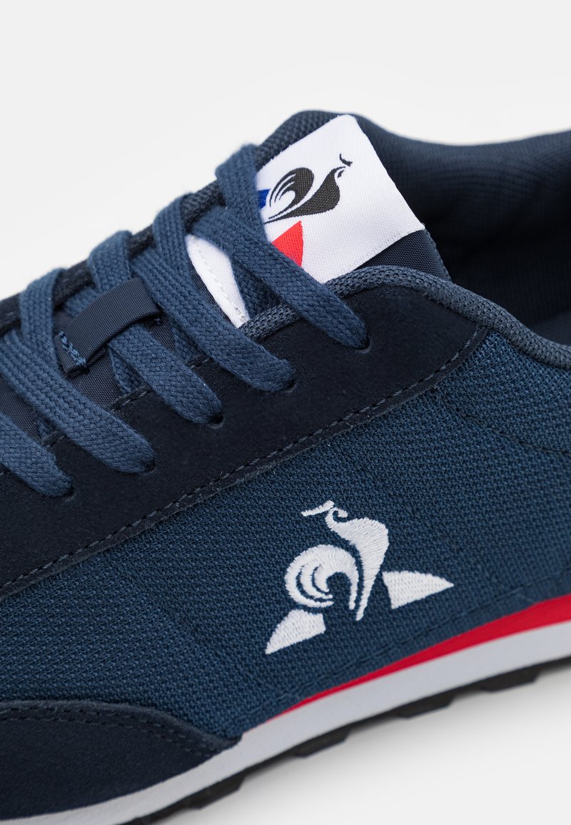 Zapatilla de deporte azul marino con materiales de tela y ante, con un logo blanco y acentos rojos en la suela. Los cordones son azul marino con un acabado texturizado.