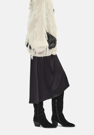 Osoba ubrana w biały sweter z frędzlami, czarną spódnicę midi, czarne zamszowe kozaki za kolano, trzyma małą czarną kopertówkę.