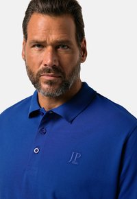 Sininen polo-paita, joka on valmistettu hengittävästä kankaasta. Tässä paidassa on klassinen kaulus, kaksinappinen avaus ja hienovarainen logo-embroidointi rinnassa.