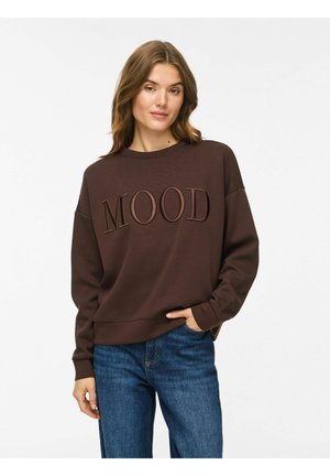 Femme aux cheveux châtain clair porte un sweat-shirt marron foncé avec le texte "MOOD" et un jean bleu, debout devant un fond clair uni.