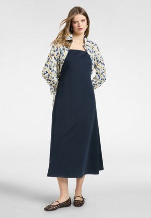 Donna che indossa un abito midi senza spalline blu navy con una camicia aperta a fantasia sopra e ballerine intrecciate marroni, in piedi davanti a uno sfondo neutro.