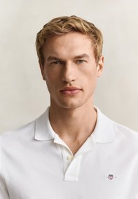 Jeune homme aux cheveux blonds et aux yeux bleus portant un polo blanc à col sur un fond uni clair.
