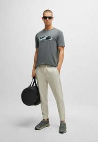 T-shirt gris à manches courtes avec un design abstrait, pantalon beige clair à cordon, baskets grises et un sac rond noir. Texture de tissu lisse.